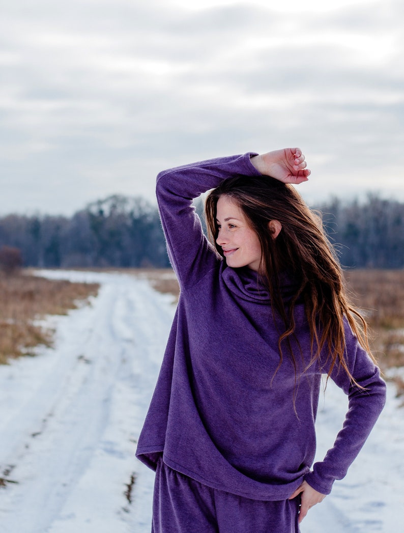May include: A woman wearing a purple long-sleeved top and matching pants stands in a snowy field. She is looking off to the side and has her hand on her head.