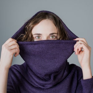 May include: A woman wearing a purple cowl neck sweater with the hood pulled up over her head. She is looking at the camera with her eyes peeking out from under the hood.