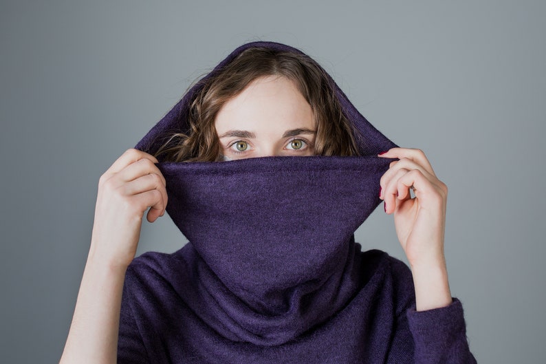 May include: A woman wearing a purple cowl neck sweater with the hood pulled up over her head. She is looking at the camera with her eyes peeking out from under the hood.