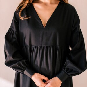 May include: A black dress featuring a V-neckline and long, voluminous sleeves. The dress has a tiered skirt and is made of a smooth material. The model's hands are clasped in front of the dress.