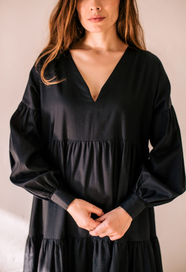 May include: A black dress featuring a V-neckline and long, voluminous sleeves. The dress has a tiered skirt and is made of a smooth material. The model's hands are clasped in front of the dress.