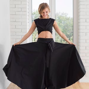 May include: A woman wearing a black crop top and wide-legged pants with a tie belt. The pants are flowing and billowing.