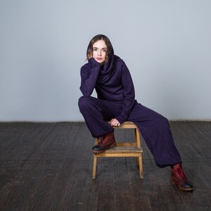 May include: A person wearing a purple hooded jumpsuit sits on a wooden stool. The person is looking at the camera with a thoughtful expression. The background is a plain white wall.