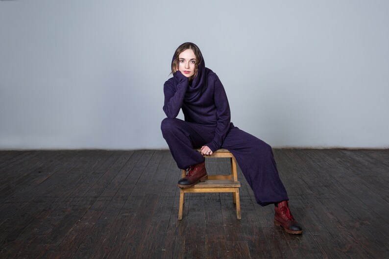 May include: A person wearing a purple hooded jumpsuit sits on a wooden stool. The person is looking at the camera with a thoughtful expression. The background is a plain white wall.