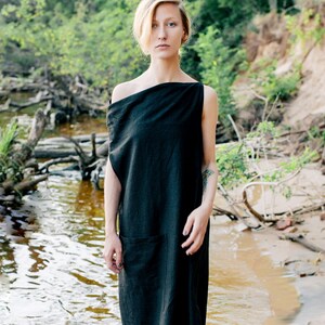 May include: A woman with blonde hair stands in a riverbed wearing a black, one-shoulder dress. The dress has a simple, loose fit and a pocket on the left side. The background is a blurred image of a river and trees.