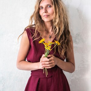 May include: A woman with long, wavy blonde hair is wearing a burgundy sleeveless jumpsuit with a sash tied at the waist. She is holding a bouquet of yellow flowers.