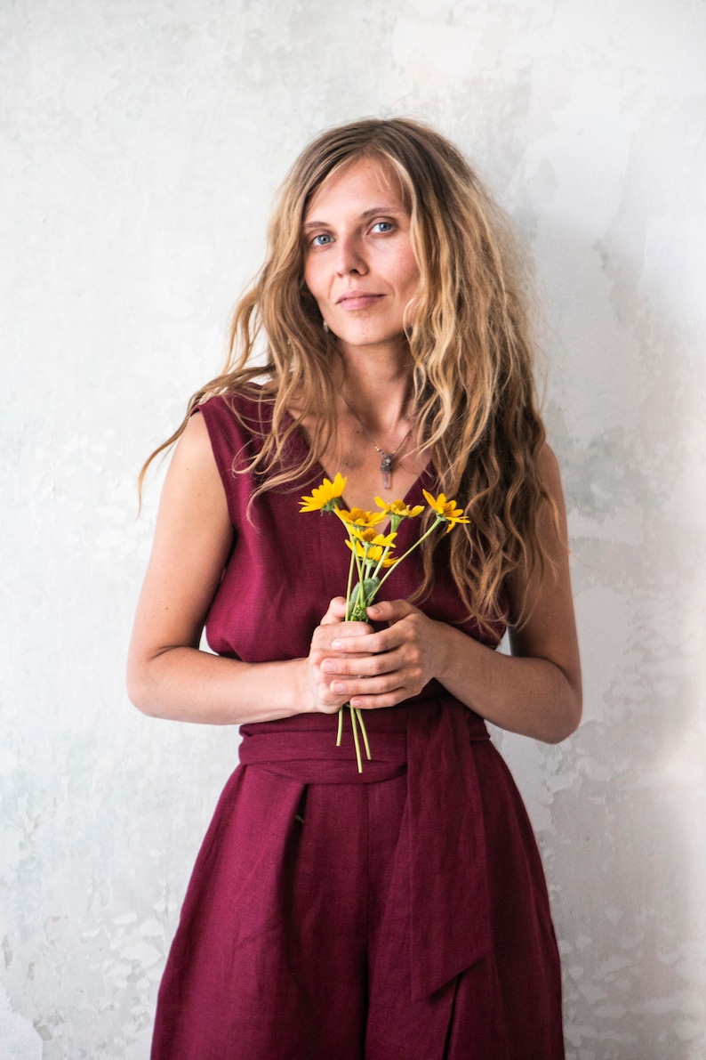 May include: A woman with long, wavy blonde hair is wearing a burgundy sleeveless jumpsuit with a sash tied at the waist. She is holding a bouquet of yellow flowers.