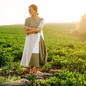 Könnte beinhalten: Eine Frau in einem langen, fließenden, grün-weißen Kleid steht auf einem Feld mit grünen Pflanzen. Die Sonne scheint hell im Hintergrund.