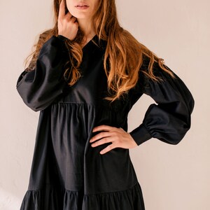 May include: A black dress featuring a V-neck and long sleeves with gathered cuffs. The dress has a tiered skirt and a relaxed silhouette. The model is wearing the dress. The dress is made of a soft, flowing fabric.