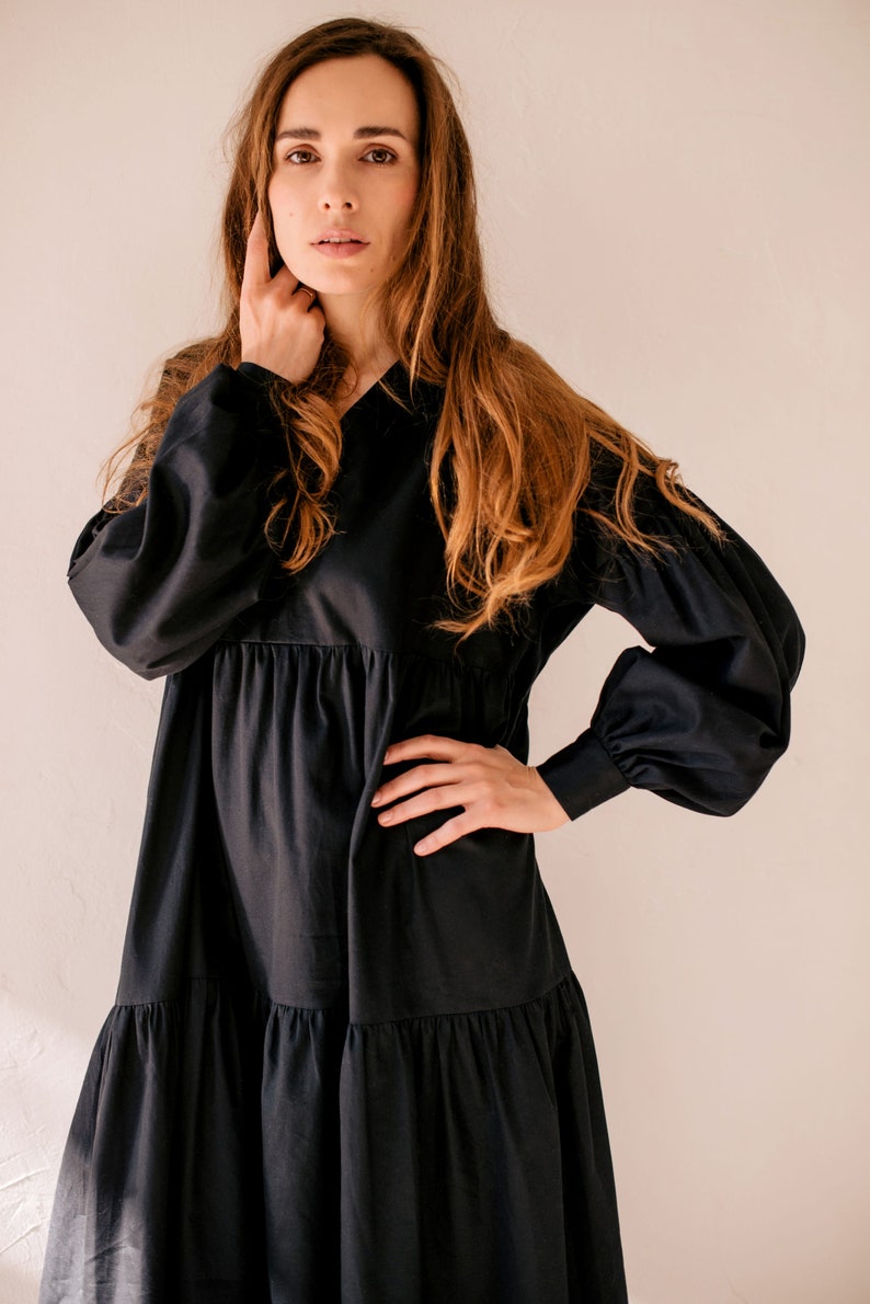 May include: A black dress featuring a V-neck and long sleeves with gathered cuffs. The dress has a tiered skirt and a relaxed silhouette. The model is wearing the dress. The dress is made of a soft, flowing fabric.
