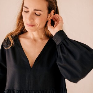 May include: A black dress featuring a deep V-neck and long sleeves with buttoned cuffs. The dress has a tiered skirt. The woman in the image is touching her ear. The dress is made of a soft fabric.