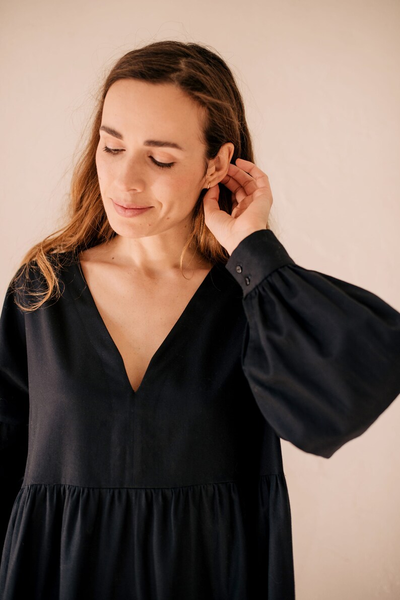 May include: A black dress featuring a deep V-neck and long sleeves with buttoned cuffs. The dress has a tiered skirt. The woman in the image is touching her ear. The dress is made of a soft fabric.