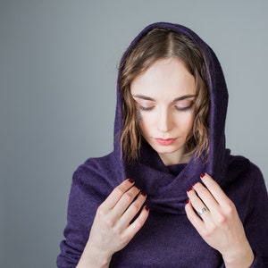 May include: A woman wearing a purple hooded sweater with a cowl neck. She is looking down and adjusting the hood with her hands.