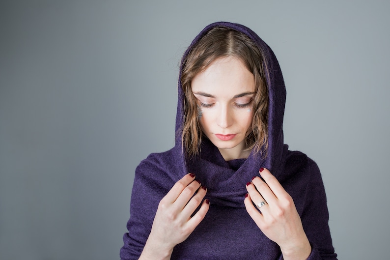 May include: A woman wearing a purple hooded sweater with a cowl neck. She is looking down and adjusting the hood with her hands.