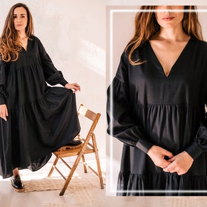 May include: A black maxi dress featuring a V-neck and long, voluminous sleeves. The dress has a tiered skirt and is paired with black shoes. The model is standing on a wooden chair in a bright room.