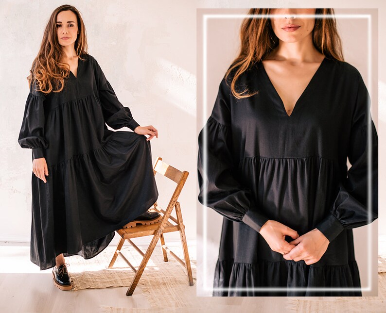 May include: A black maxi dress featuring a V-neck and long, voluminous sleeves. The dress has a tiered skirt and is paired with black shoes. The model is standing on a wooden chair in a bright room.