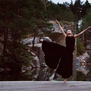 May include: A woman in a black dress dances on a wooden platform in front of a rocky landscape and a body of water.