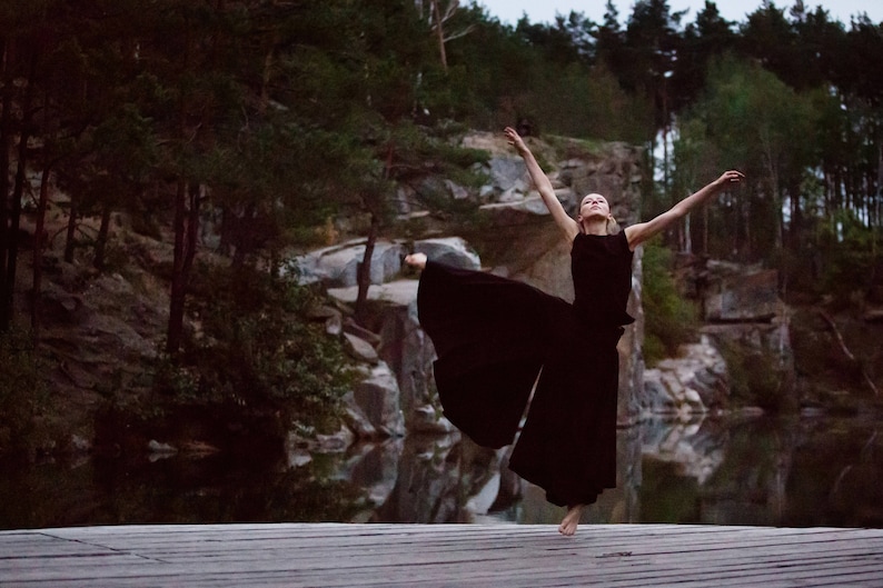 May include: A woman in a black dress dances on a wooden platform in front of a rocky landscape and a body of water.