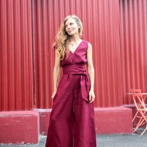May include: A woman wearing a burgundy wide-leg jumpsuit with a sash tie at the waist. She is standing in front of a red corrugated metal wall.