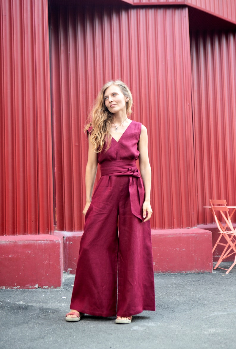 May include: A woman wearing a burgundy wide-leg jumpsuit with a sash tie at the waist. She is standing in front of a red corrugated metal wall.