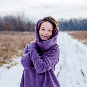 May include: A woman wearing a purple hooded sweater stands in a snowy field. She is smiling and looking at the camera.