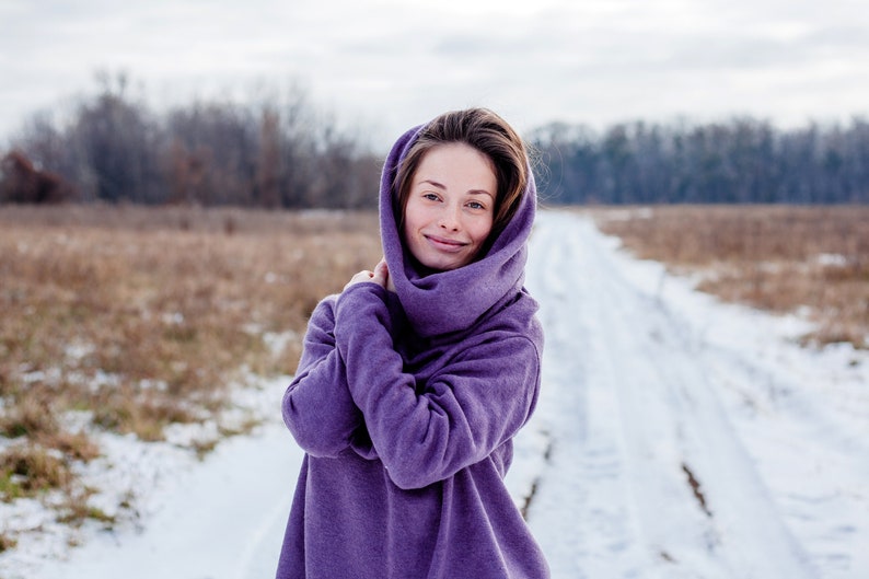 May include: A woman wearing a purple hooded sweater stands in a snowy field. She is smiling and looking at the camera.