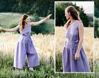 May include: A woman wearing a lavender linen jumpsuit with a tie at the waist. The jumpsuit has a wide leg and a sleeveless design. The woman is standing in a field of tall grass.
