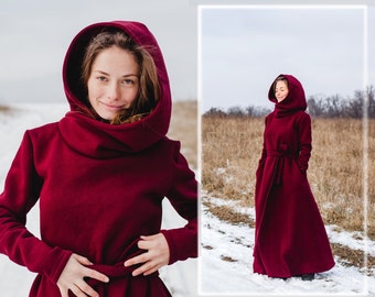 May include: A woman wearing a long, burgundy hooded sweater dress with a high collar and a tie belt. The dress is made of a soft, warm fabric and has a relaxed fit. The woman is standing in a snowy field with a blurred background.