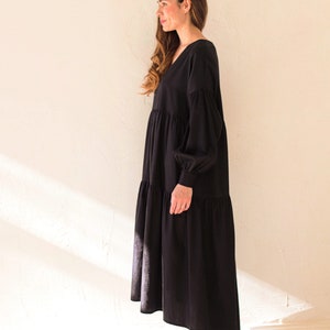 May include: A black maxi dress with a V-neck and long, voluminous sleeves. The dress has a tiered skirt and is made of a soft fabric. The model is wearing black leather shoes.