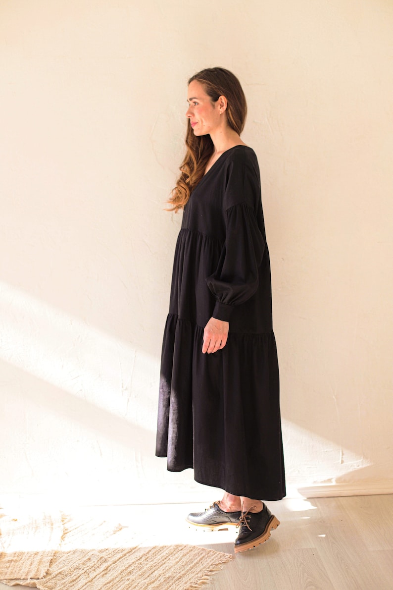 May include: A black maxi dress with a V-neck and long, voluminous sleeves. The dress has a tiered skirt and is made of a soft fabric. The model is wearing black leather shoes.