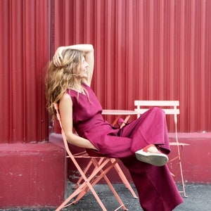 May include: A woman wearing a burgundy jumpsuit sits on a pink folding chair in front of a red corrugated metal wall.