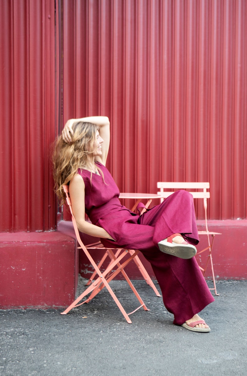 May include: A woman wearing a burgundy jumpsuit sits on a pink folding chair in front of a red corrugated metal wall.