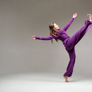 May include: A woman in a purple jumpsuit performs a dance move with her leg extended high in the air.