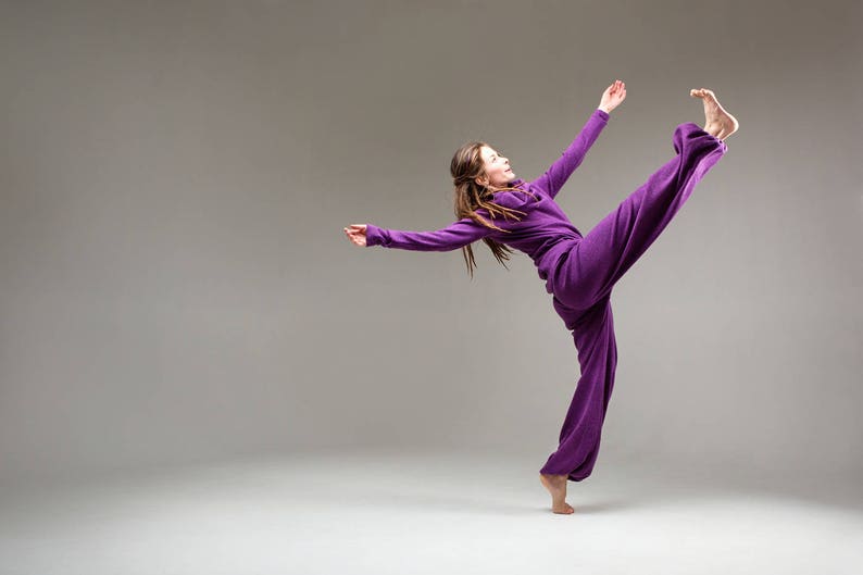 May include: A woman in a purple jumpsuit performs a dance move with her leg extended high in the air.