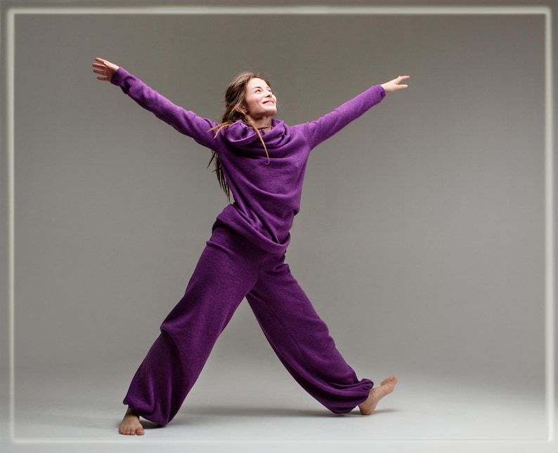 May include: A woman wearing a purple, long-sleeved jumpsuit with a cowl neck. She is standing with her arms outstretched and her head tilted back, looking up.