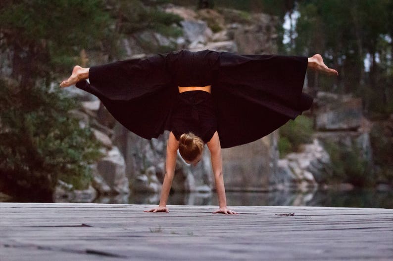 May include: A person in black clothing performs a handstand on a wooden surface. The person's legs are extended and their arms are straight. The background is a rocky, outdoor setting.