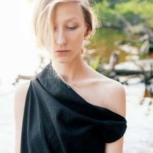May include: A woman with blonde hair wearing a black one-shoulder top. She is standing in front of a blurred background of water and greenery.