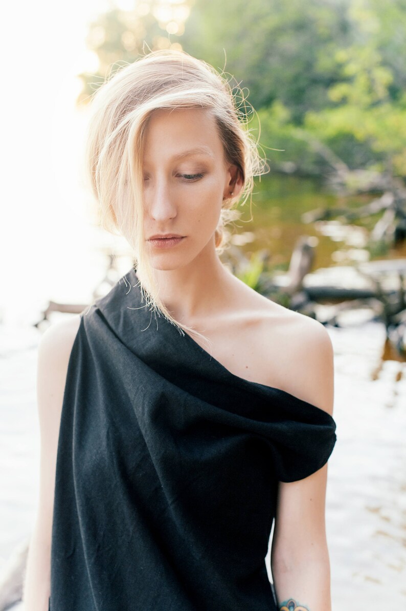 May include: A woman with blonde hair wearing a black one-shoulder top. She is standing in front of a blurred background of water and greenery.