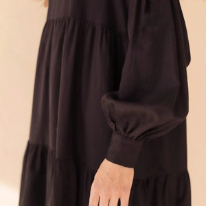May include: Black tiered dress with long, voluminous sleeves. The dress features a V-neckline and a relaxed fit. The sleeves have gathered details at the shoulders and cuffs. The dress is made of a soft, flowing fabric.