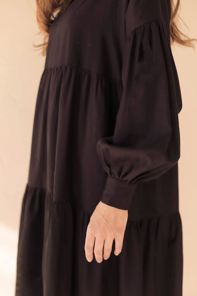 May include: Black tiered dress with long, voluminous sleeves. The dress features a V-neckline and a relaxed fit. The sleeves have gathered details at the shoulders and cuffs. The dress is made of a soft, flowing fabric.
