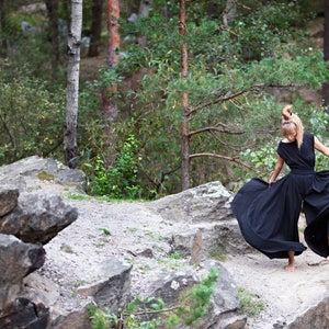 May include: A woman wearing a black maxi dress with a flowing skirt stands on a rocky path in a forest. The dress has a simple design and is made of a lightweight fabric.