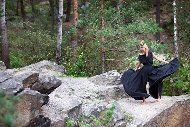 May include: A woman wearing a black maxi dress with a flowing skirt stands on a rocky path in a forest. The dress has a simple design and is made of a lightweight fabric.