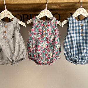 May include: Three baby onesies hanging on wooden hangers. The onesies are made of cotton and have different patterns: one with a gray background and white animal prints, one with a floral print, and one with a blue and white gingham pattern.