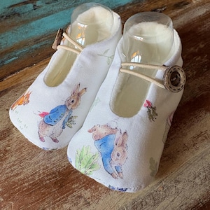 Könnte beinhalten: Ein Paar Babyschuhe aus weißem Stoff mit einem Druckmuster von Peter Hase in einer blauen Jacke, umgeben von kleinen grünen Gemüsesorten. Jeder Schuh hat einen beigen Gummizug, der mit einem runden braunen Holzknopf befestigt ist.