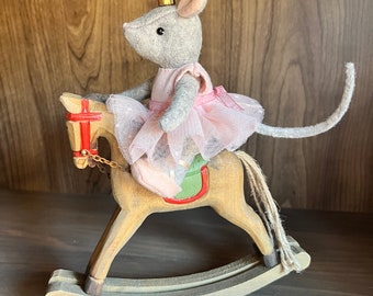 May include: A gray mouse wearing a pink tutu and a gold crown rides a wooden rocking horse. The horse has a red saddle and a green and brown design.
