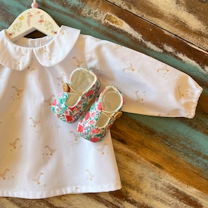 Puede incluir: Una blusa blanca para bebé con cuello de Peter Pan y un estampado de gansos. La blusa está sobre una superficie de madera con un par de zapatos de bebé con estampado floral.