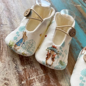 May include: A pair of baby shoes with a Peter Rabbit print. The shoes are white with a blue and green pattern. The shoes have a brown button on each shoe.