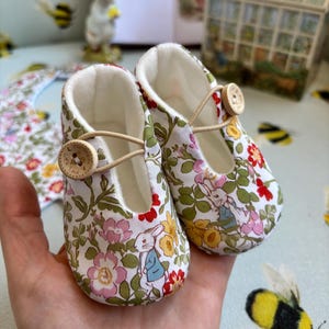 Può includere: Un paio di scarpine per neonati con stampa floreale che presenta un design di coniglio. Le scarpine sono bianche con fiori rosa, rossi, gialli e verdi. Le scarpine hanno una chiusura a bottone marrone e una fodera bianca.