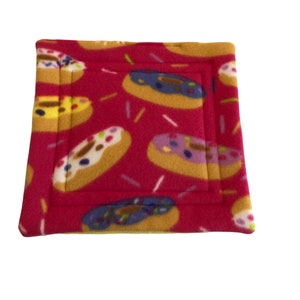Peut inclure: Un coussin pour animaux de compagnie en polaire rouge avec un motif répété de beignets avec différents glaçages et vermicelles colorés. Les beignets ont un glaçage jaune, bleu, violet et blanc. Le lit a une bordure cousue.