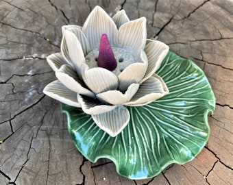 May include: A white ceramic lotus flower incense burner with a green leaf base. The incense cone is purple and sits in the center of the flower.